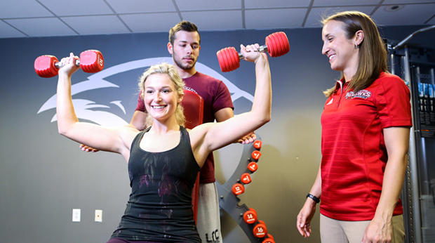 A La Roche University student lifts dumbbells in a weight room. A student stands behind her to help with her form. An instructor stands next to them. 